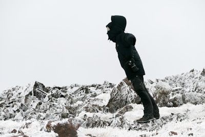A person dressed in heavy winter clothing stands on a rocky, snow-covered terrain. They are wearing a hooded jacket and boots, appearing to brace against the cold, with a small camera hanging by their side. The landscape is dominated by rugged rocks and a light dusting of snow, under a gray sky.