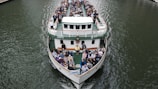 A large ferry is filled with passengers seated on its open deck, navigating through a body of water. People are seated facing forward, with a clear view of the waterway. The boat features multiple levels and is designed for river cruises or tours. The water appears calm and the boat is cruising steadily, creating gentle ripples as it moves.