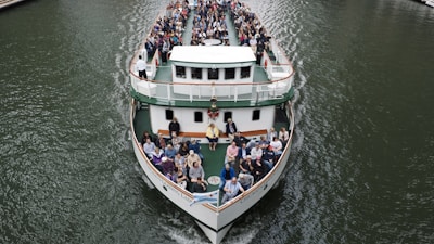 A large ferry is filled with passengers seated on its open deck, navigating through a body of water. People are seated facing forward, with a clear view of the waterway. The boat features multiple levels and is designed for river cruises or tours. The water appears calm and the boat is cruising steadily, creating gentle ripples as it moves.