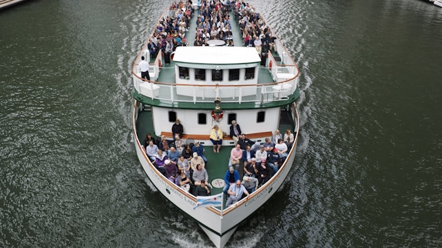 A large ferry is filled with passengers seated on its open deck, navigating through a body of water. People are seated facing forward, with a clear view of the waterway. The boat features multiple levels and is designed for river cruises or tours. The water appears calm and the boat is cruising steadily, creating gentle ripples as it moves.