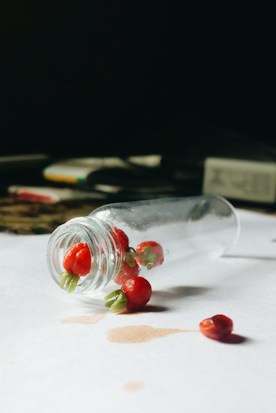 A small glass bottle lies on its side with red berries spilling out onto a white surface. The berries are small, smooth, and vivid red with green stems, creating a striking contrast against the glass. A soft shadow is cast around the bottle and berries, with some liquid marks on the paper adding to the natural, casual appearance. In the blurred background, indistinct objects suggest a dimly lit environment.