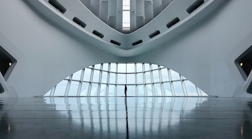 A modern and minimalistic interior with a futuristic architectural design. The space features curved lines and large windows that allow natural light to illuminate the area. The reflection on the glossy floor adds depth to the scene.