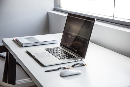 A modern laptop with a reflective screen sits on a minimalist white desk. Nearby, there is a stack of closed notebooks or planners and a wireless mouse. A pair of glasses lies nearby. The setting appears well-lit due to natural light streaming through a large window.