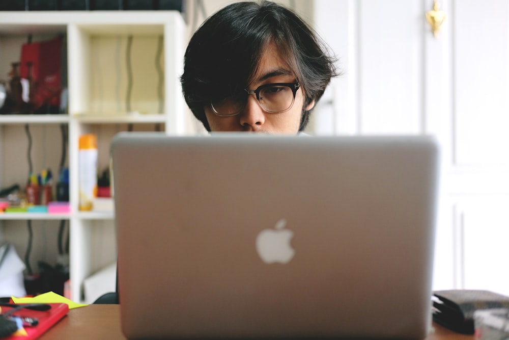 man wearing eyeglasses while using silver Macbook near wooden cubby shelf inside room