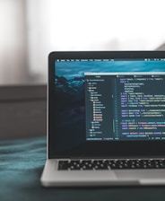 A laptop displaying a code editor with lines of code related to a React application. The screen shows project folders and files written in JavaScript, suggesting software development in progress. The background is slightly blurred, adding focus to the laptop screen.