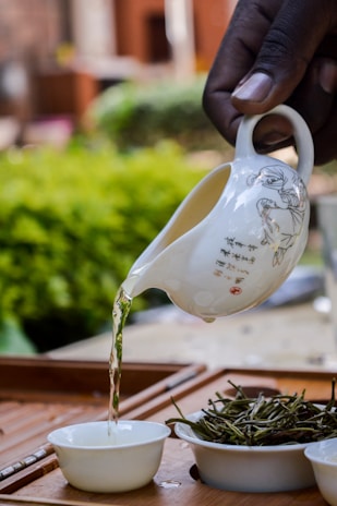 A hand is pouring liquid from a decorative white ceramic pitcher into a small white bowl. The pitcher has intricate designs and some text. Next to the bowl, there is a pile of dried green leaves. The background features blurred greenery and a wooden surface.