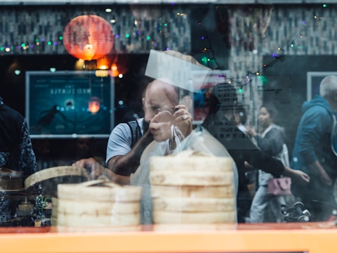 A chef is seen through a window reflection as they prepare food, with bamboo steamers prominently displayed. The scene is lively with people walking by outside and colorful lights hanging above. A red lantern adds a traditional touch to the setting.