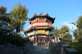 A traditional multi-tiered Chinese pavilion with intricate wooden carvings and red accents stands prominently at the end of a stone staircase. The structure is surrounded by lush green trees and shrubs under a clear blue sky. Decorative signage with Chinese characters is visible above the entrance.