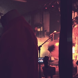 A dimly lit stage environment featuring a musician in motion, with a microphone stand in the foreground. Atmospheric lighting creates an intimate mood, and a camera on a tripod captures the scene.