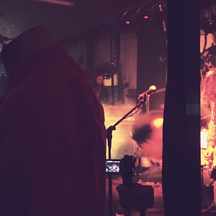 A dimly lit stage environment featuring a musician in motion, with a microphone stand in the foreground. Atmospheric lighting creates an intimate mood, and a camera on a tripod captures the scene.