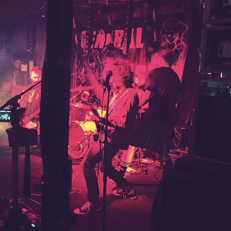 A band performs energetically on stage, surrounded by musical equipment and vibrant stage lighting. The background features a banner with partial text and abstract designs. The atmosphere is lively with musicians engaged in playing instruments.