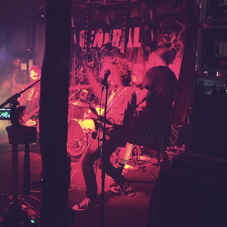 A band performs energetically on stage, surrounded by musical equipment and vibrant stage lighting. The background features a banner with partial text and abstract designs. The atmosphere is lively with musicians engaged in playing instruments.