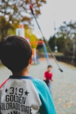 A young child wearing a sweatshirt with the text '319 Golden Colorado State' stands in the foreground, looking towards another child on a playground setting. The background consists of outdoor playground structures and trees with autumn foliage.
