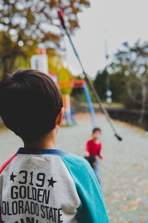 A young child wearing a sweatshirt with the text '319 Golden Colorado State' stands in the foreground, looking towards another child on a playground setting. The background consists of outdoor playground structures and trees with autumn foliage.