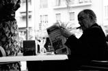 An elderly man with a cigar is sitting at a cafe table, reading a newspaper. A glass of wine and some tableware are placed on the table. The background displays city buildings and a tree, suggesting an outdoor urban setting. The image is in black and white.