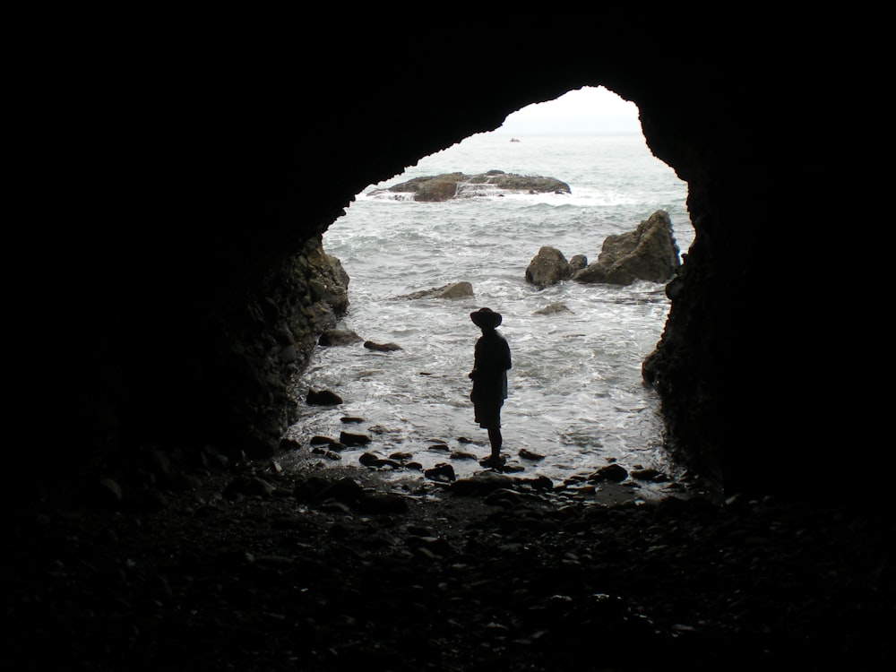 silhouette of person wearing hat taking picture inside cave near sea water