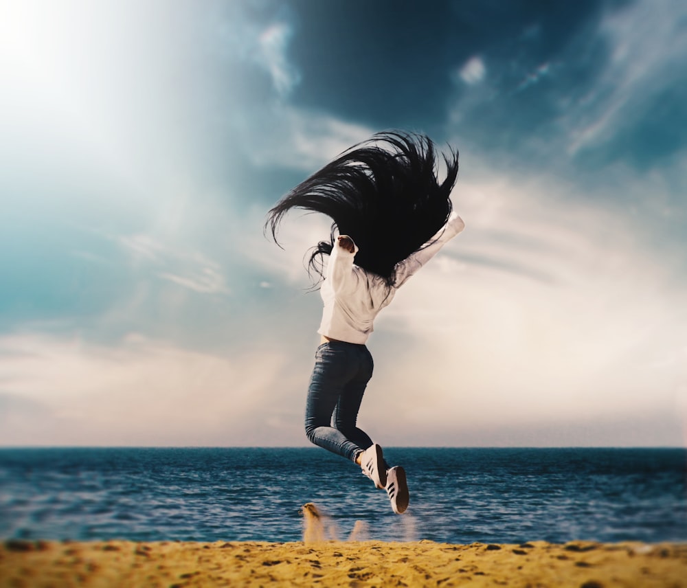 woman in white long-sleeved shirt and blue jeans jumping on seashore during daytime