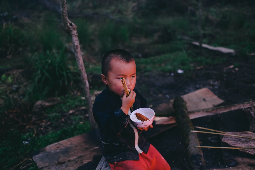 boy earring food while holding round white bowl with pair of chopsticks