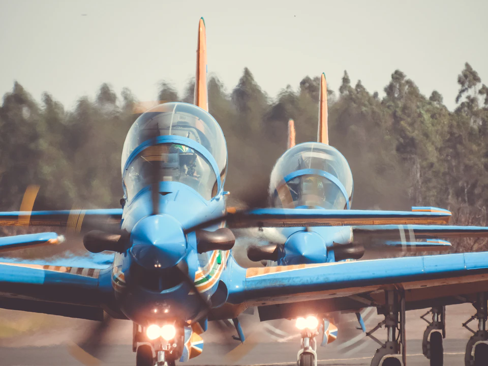 Two blue and yellow airplanes stand side by side on a runway with spinning propellers