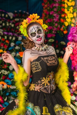 A person is dressed in a traditional costume with intricate face paint resembling a skull, embodying the Day of the Dead theme. The outfit features vibrant patterns and embroidery, complemented by a colorful headdress with flowers. The background is adorned with vivid, multicolored flowers, enhancing the festive and cultural atmosphere.