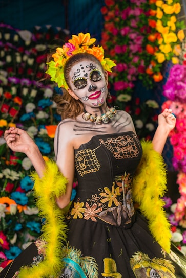 A person is dressed in a traditional costume with intricate face paint resembling a skull, embodying the Day of the Dead theme. The outfit features vibrant patterns and embroidery, complemented by a colorful headdress with flowers. The background is adorned with vivid, multicolored flowers, enhancing the festive and cultural atmosphere.