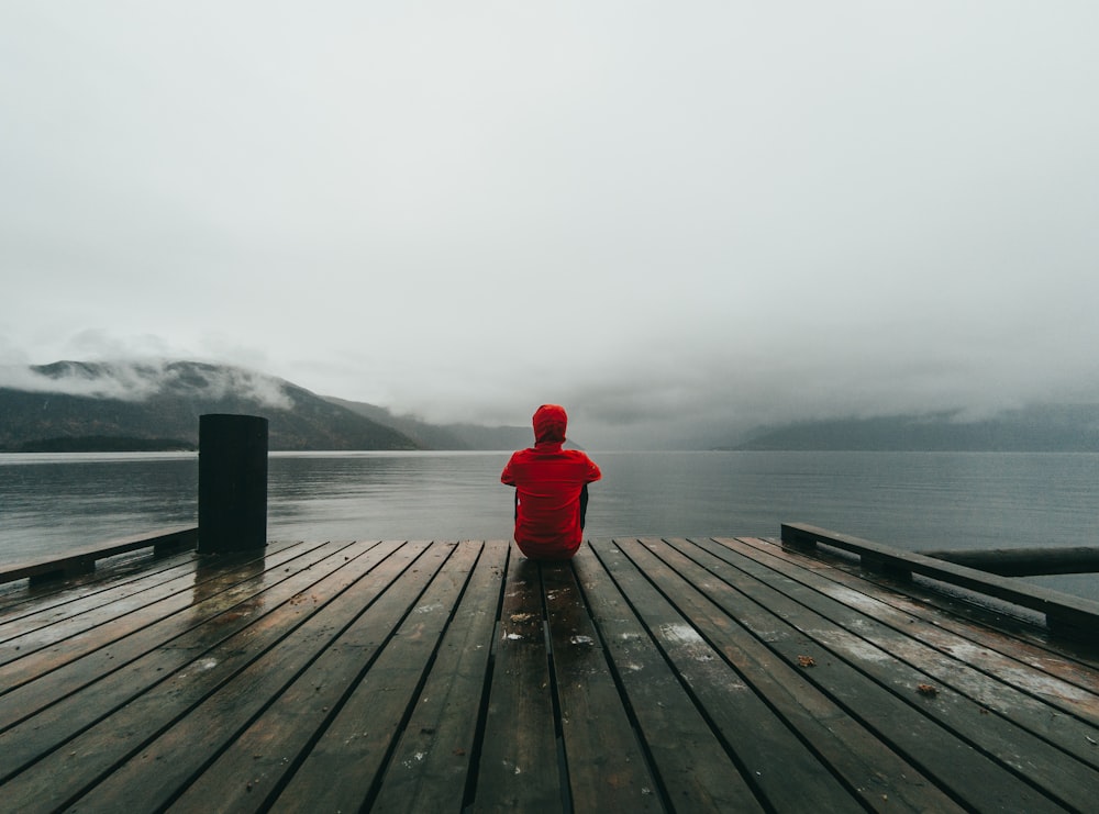 person wearing red hooded jacket sitting on brown and black wooden sea dock near mountains under gray sky during daytime