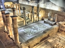 An ancient archaeological site with tall stone columns and intricate mosaic flooring. The area is illuminated by soft lighting, highlighting the architectural details and artwork. The mosaics depict various animals and mythological figures, surrounded by geometric patterns. The setting is enclosed by solid stone walls, and historical artifacts are displayed within the space.