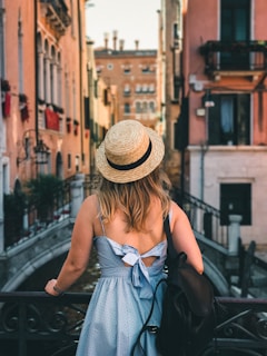 A woman with long hair stands on a bridge, looking toward historic European architecture with narrow buildings lining a canal. She wears a straw hat and a light blue dress with a bow on the back, and holds a black backpack.