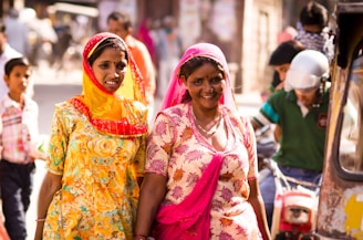 Two women dressed in vibrant traditional clothing walk side by side on a busy street. The woman on the left wears a bright yellow dress with floral patterns and a red-tinged scarf, while the woman on the right wears a pink and white patterned dress with a pink scarf. Background elements include other people, a man on a motorbike wearing a helmet, and blurred street activities.