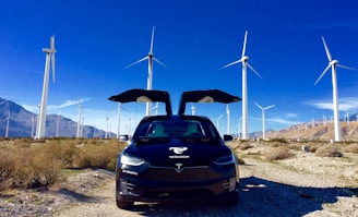 A black Tesla car with its falcon-wing doors open is parked in a desert landscape. Surrounding the car are numerous tall wind turbines, scattered across the dry, shrub-filled terrain. In the background, there are distant mountain ranges under a vibrant, clear blue sky.