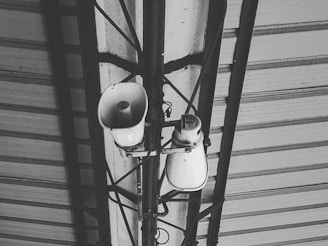 A black and white image featuring a metal roof structure with two loudspeakers mounted on a metal frame. Various cables are visible connecting to the speakers, which appear to be used for public announcements.