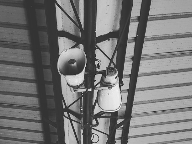 A black and white image featuring a metal roof structure with two loudspeakers mounted on a metal frame. Various cables are visible connecting to the speakers, which appear to be used for public announcements.