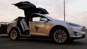 A white Tesla vehicle is pictured with its falcon-wing doors open, displaying a modern and sleek interior under a twilight sky. The car is parked on a concrete surface, visible in profile with branding on the door.