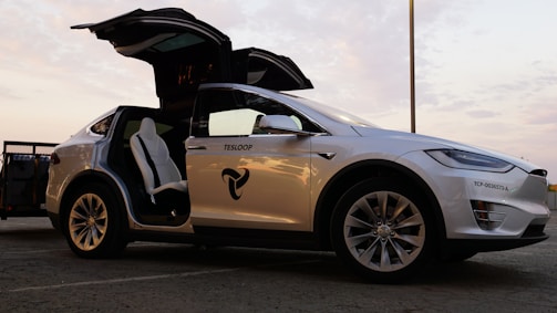 A white Tesla vehicle is pictured with its falcon-wing doors open, displaying a modern and sleek interior under a twilight sky. The car is parked on a concrete surface, visible in profile with branding on the door.