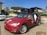 A red Tesla with its falcon-wing doors open is parked on a sunny residential street. The car has the word 'TESLOOP' and a logo on the side and front. Nearby is another car, and there are houses surrounded by greenery. The sky is clear and blue.