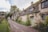 A row of charming stone cottages with steeply pitched roofs, covered with moss and vines. The cottages are set along a narrow road bordered by lush greenery. In the background, several large trees and a pathway with a few people walking create a tranquil, picturesque village atmosphere.