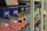A close-up view of shelves filled with various cans of paint, with a focus on colorful labels, including pink, orange, and blue hues. The depth of field blurs the background, highlighting the foreground cans.