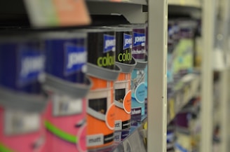 A close-up view of shelves filled with various cans of paint, with a focus on colorful labels, including pink, orange, and blue hues. The depth of field blurs the background, highlighting the foreground cans.