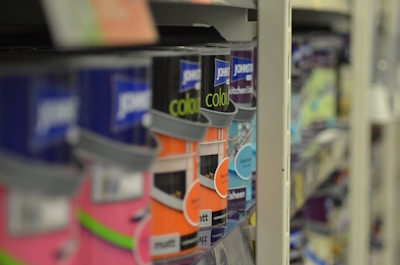 A close-up view of shelves filled with various cans of paint, with a focus on colorful labels, including pink, orange, and blue hues. The depth of field blurs the background, highlighting the foreground cans.
