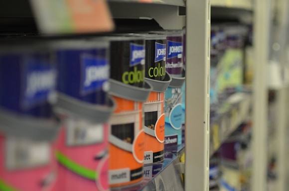 A close-up view of shelves filled with various cans of paint, with a focus on colorful labels, including pink, orange, and blue hues. The depth of field blurs the background, highlighting the foreground cans.
