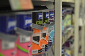 A close-up view of shelves filled with various cans of paint, with a focus on colorful labels, including pink, orange, and blue hues. The depth of field blurs the background, highlighting the foreground cans.