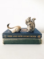Two vintage books are stacked on top of each other with decorative metallic sculptures of a squirrel and a whale tail placed on the top book. The books have worn covers, one blue titled 'HERE AND NOW STORY BOOK' and the other green, without visible titling. The sculptures suggest a whimsical or playful theme.