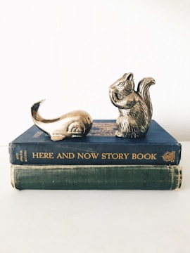 Two vintage books are stacked on top of each other with decorative metallic sculptures of a squirrel and a whale tail placed on the top book. The books have worn covers, one blue titled 'HERE AND NOW STORY BOOK' and the other green, without visible titling. The sculptures suggest a whimsical or playful theme.