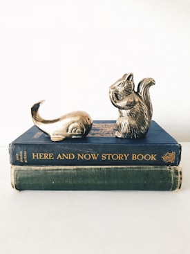 Two vintage books are stacked on top of each other with decorative metallic sculptures of a squirrel and a whale tail placed on the top book. The books have worn covers, one blue titled 'HERE AND NOW STORY BOOK' and the other green, without visible titling. The sculptures suggest a whimsical or playful theme.