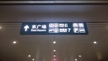 A ceiling-mounted directional sign in a public transportation or airport facility. It displays bilingual text in Chinese and English, with icons indicating directions to the East Square, arrival gates A1 and B1, a ticket office, a parking area, and a bus station.