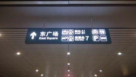 A ceiling-mounted directional sign in a public transportation or airport facility. It displays bilingual text in Chinese and English, with icons indicating directions to the East Square, arrival gates A1 and B1, a ticket office, a parking area, and a bus station.