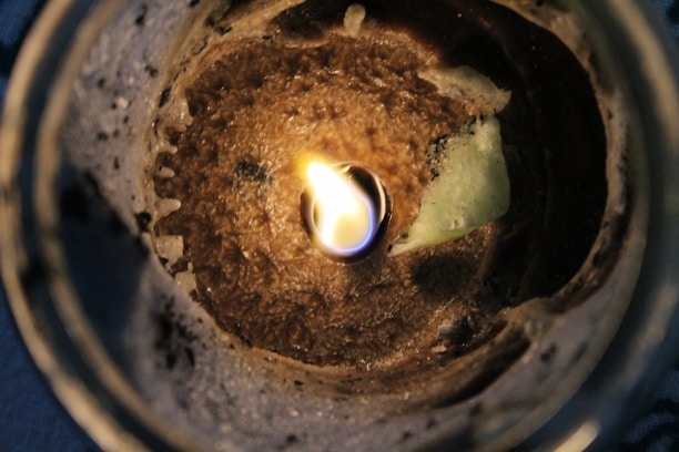 A close-up view of a candle with partially melted wax and a small flame burning in the center. The wax appears to be a light brown color, and there is an uneven texture around the edges of the wax. The flame casts a warm glow that illuminates the surrounding area.