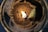 A close-up view of a candle with partially melted wax and a small flame burning in the center. The wax appears to be a light brown color, and there is an uneven texture around the edges of the wax. The flame casts a warm glow that illuminates the surrounding area.