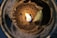 A close-up view of a candle with partially melted wax and a small flame burning in the center. The wax appears to be a light brown color, and there is an uneven texture around the edges of the wax. The flame casts a warm glow that illuminates the surrounding area.
