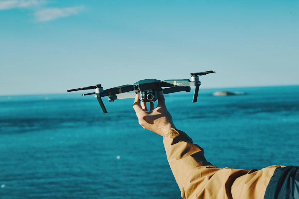 person holding grey drone in front of sea